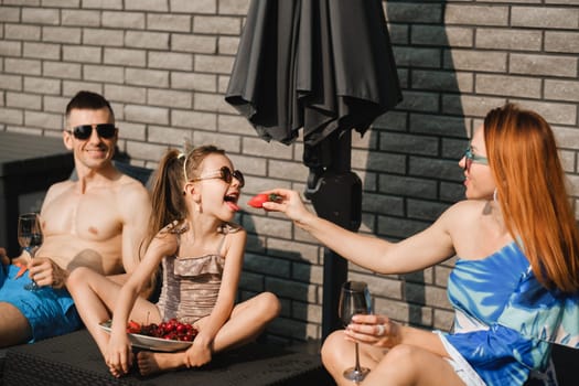 A happy family in swimsuits sunbathes on their terrace in summer. Mom feeds her daughter strawberries.