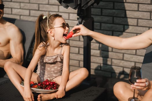 A happy family in swimsuits sunbathes on their terrace in summer. Mom feeds her daughter strawberries.