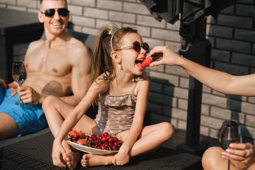 A happy family in swimsuits sunbathes on their terrace in summer. Mom feeds her daughter strawberries.