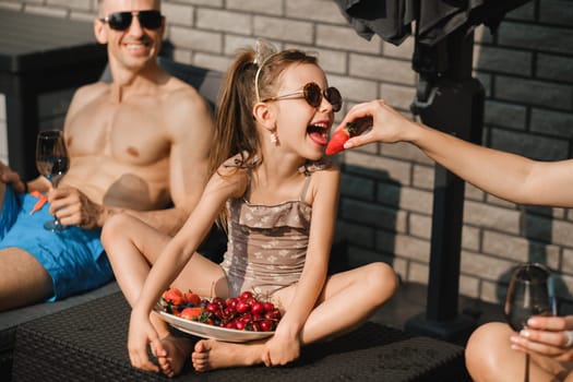 A happy family in swimsuits sunbathes on their terrace in summer. Mom feeds her daughter strawberries.