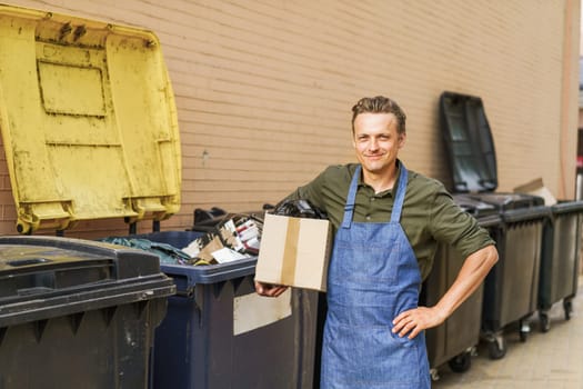 Garbage sorting with blond smiling man wearing blue apron. Man holds box filled with garbage near city trash can in back yard. Importance of responsible waste management and recycling practices. . High quality photo