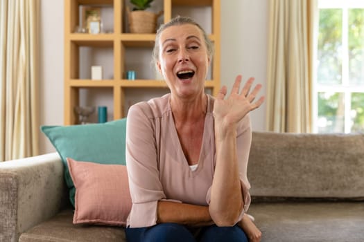 Caucasian senior woman laughing and waving hand while sitting on sofa at home. unaltered, lifestyle, retirement, leisure, happiness, domestic life.