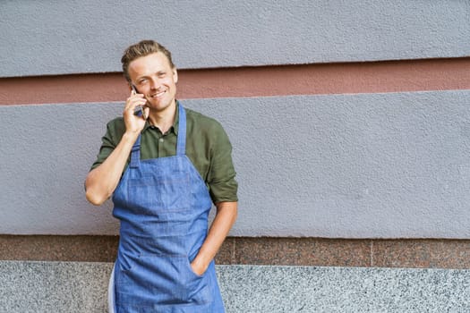 Concept of service readiness and the willingness to assist customers. Worker in blue apron, holding mobile phone, and ready to take order from client against wall background. High quality photo