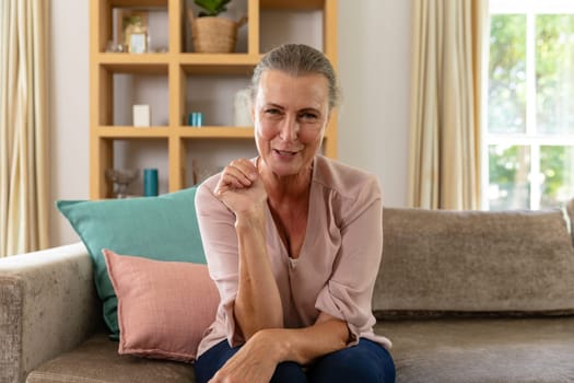 Portrait of smiling caucasian senior woman sitting on sofa at home. unaltered, lifestyle, retirement, leisure, domestic life.