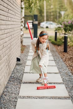 A little girl with a brush cleans a path on the street in the courtyard.