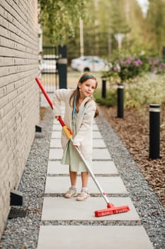 A little girl with a brush cleans a path on the street in the courtyard.