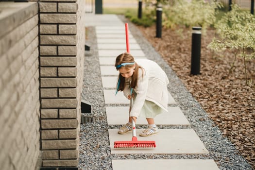 A little girl with a brush cleans a path on the street in the courtyard.