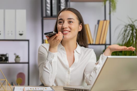 Caucasian young business woman working, having mobile phone loudspeaker talk at office desk with laptop. Manager freelance girl holding smartphone using messenger chat apps. Employment, occupation