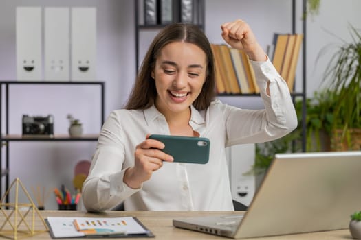 Young business woman playing on smartphone exciting mobile application celebrate winning racing, shooter game at office desk workplace. Professional manager freelancer girl enjoy good work of new app