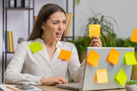 Tired exhausted young business woman working on laptop at office with many sticker task inscriptions. Freelancer girl busy occupation feels sad boredom overworked multitasking. Panic attack, deadline