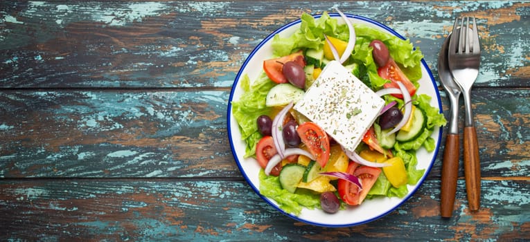 Traditional Greek Salad with Feta Cheese, Tomatoes, Bell Pepper, Cucumbers, Olives, Herbs in white ceramic bowl on blue rustic wooden table background from above, Cuisine of Greece. Space for text