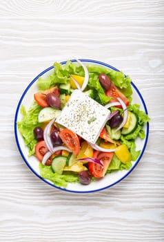 Traditional Greek Salad with Feta Cheese, Tomatoes, Bell Pepper, Cucumbers, Olives, Herbs in white ceramic bowl on White rustic wooden table background from above, Cuisine of Greece