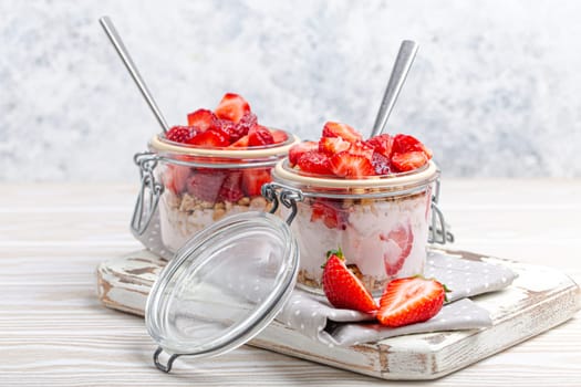 Parfait with Fresh Strawberries, Yoghurt and Crunchy Granola in Transparent Glass Mason Jars on White Rustic Wooden Background from Angle View, Healthy Breakfast or Light Summer Fruit Dessert