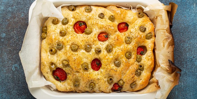 Overhead of Traditional Italian Homemade Flat Bread Focaccia with Green Olives, Olive Oil, Cherry Tomatoes and Rosemary in Baking Tray on Rustic Dark Blue Concrete Background