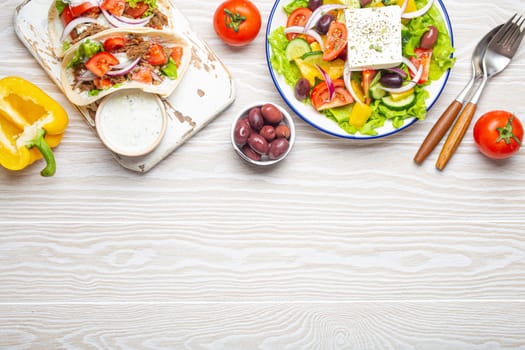 Traditional Greek Food: Greek Salad, Gyros with meat and vegetables, Tzatziki sauce, Olives on White rustic wooden table background from above. Cuisine of Greece. Copy space