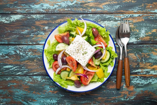 Traditional Greek Salad with Feta Cheese, Tomatoes, Bell Pepper, Cucumbers, Olives, Herbs in white ceramic bowl on blue rustic wooden table background from above, Cuisine of Greece