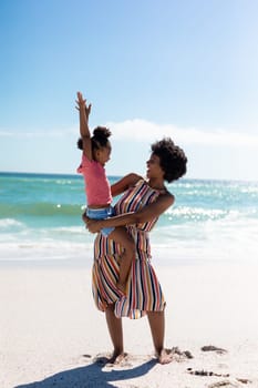 Cheerful african american mother carrying daughter while enjoying sunny day at beach against sky. unaltered, family, lifestyle, togetherness, enjoyment and holiday concept.