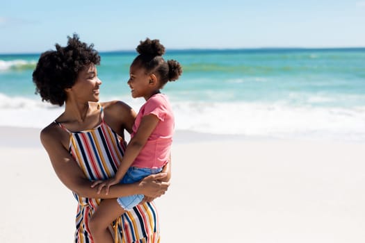 Happy african american woman carrying daughter while standing at beach on sunny day. unaltered, family, lifestyle, togetherness, enjoyment and holiday concept.