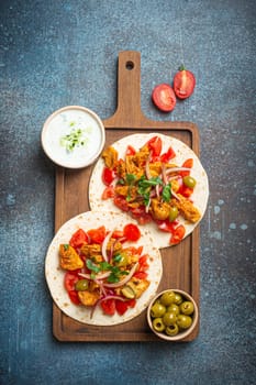 Cooking Traditional Greek Dish Gyros: Pita bread with vegetables, meat, herbs, olives on rustic wooden cutting board with Tzatziki sauce, olive oil top view on dark blue stone background.
