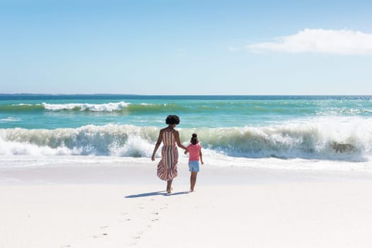 African american mother and daughter holding hands while walking towards waves at beach. unaltered, family, lifestyle, togetherness, enjoyment and holiday concept.