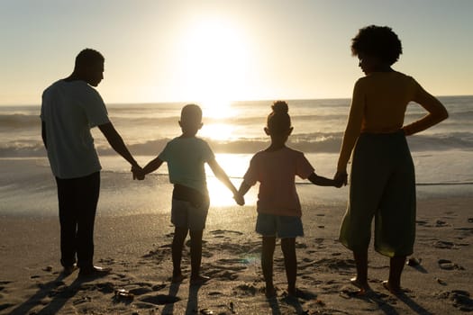 Rear view of african american family holding hands while looking at sunset over sea from beach shore. unaltered, family, lifestyle, togetherness, enjoyment and holiday concept.