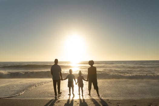 Full length rear view of african american family holding hands while looking at sunset over sea. unaltered, family, lifestyle, togetherness, enjoyment and holiday concept.