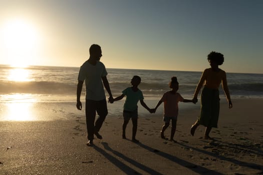 Full length of african american family holding hands walking at beach against sky during sunset. unaltered, family, lifestyle, togetherness, enjoyment and holiday concept.