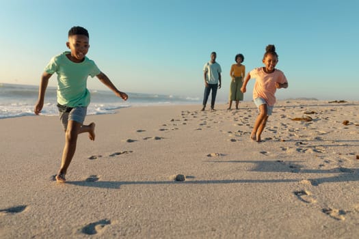 Full length of happy african american children running against parents walking at beach on sunny day. unaltered, family, lifestyle, togetherness, enjoyment and holiday concept.