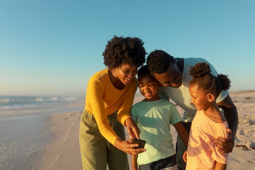 Happy african american mother sharing smartphone with family at beach on sunny day. unaltered, family, lifestyle, togetherness, technology and holiday concept.