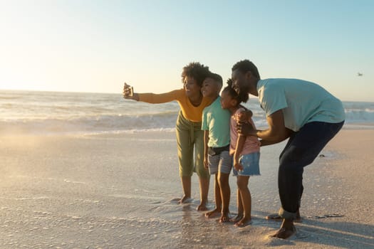 Happy african american mother taking selfie with family on shore at beach during sunset. unaltered, family, lifestyle, togetherness, technology and holiday concept.