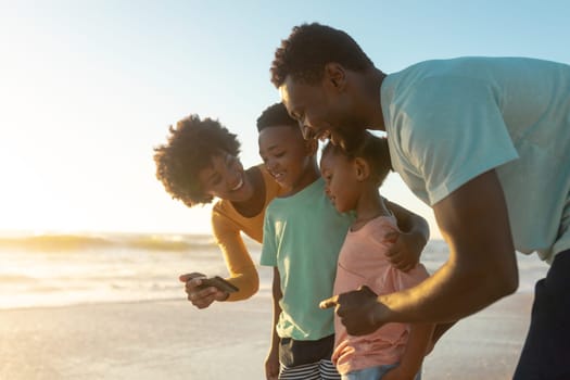 Happy african american woman sharing smartphone with family at beach during sunset. unaltered, family, lifestyle, togetherness, technology and holiday concept.