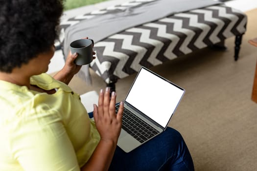 High angle view of african american mid adult woman waving hand over laptop while sitting at home. unaltered, video call, copy space, wireless technology, drink, lifestyle and domestic life concept.