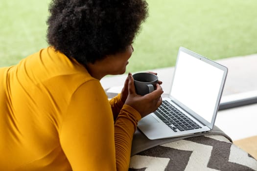 Side view of african american mid adult woman holding coffee cup while using laptop at home. unaltered, video call, wireless technology, drink, lifestyle and domestic life concept.