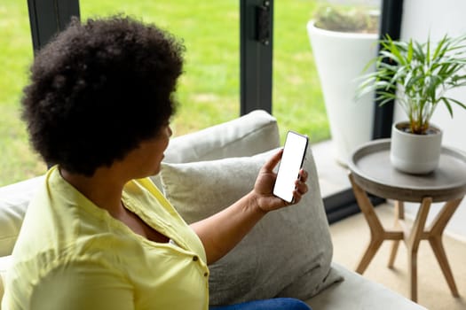 African american mid adult woman using smart phone with blank screen while sitting on sofa at home. unaltered, copy space, video call, wireless technology, lifestyle and domestic life concept.