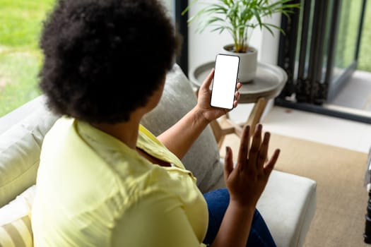 African american mid adult woman waving hand over smart phone during video call. unaltered, video call, copy space, wireless technology, drink, lifestyle and domestic life concept.