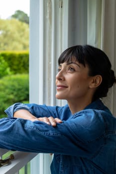 Smiling thoughtful caucasian young woman with arms crossed looking through window. unaltered, lifestyle, domestic life, home, contemplation.