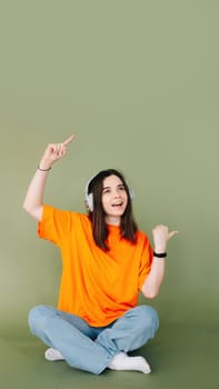Portrait of a joyful woman wearing headphones, expressing enthusiasm and approval while pointing both thumbs towards empty space, isolated on a vibrant green background.