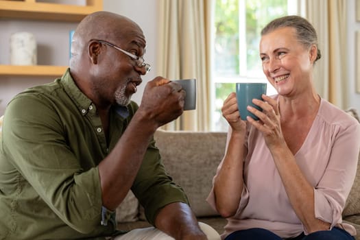 Happy senior multiracial couple having coffee while sitting on sofa at home. unaltered, lifestyle, leisure, retirement, togetherness, drink, refreshment.