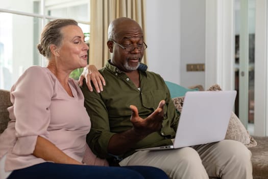 Senior multiracial couple discussing over laptop while sitting on sofa at home. unaltered, lifestyle, retirement, togetherness, leisure, wireless technology.