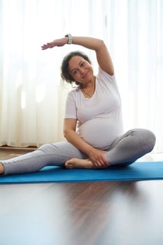 Smiling pregnant woman stretching her body while exercising on yoga mat at home. The concept of healthy active pregnancy and happy maternity leave. Prenatal fitness. Pregnancy. Maternity lifestyle