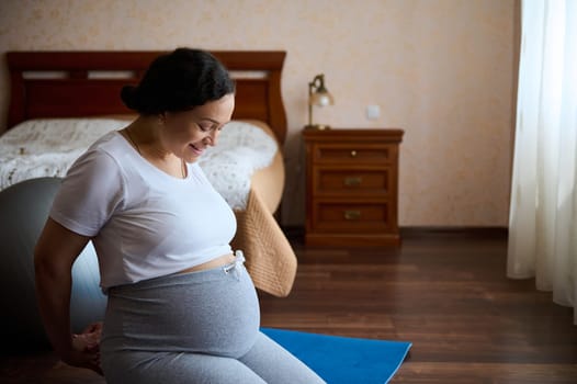 Gravid expectant mother in last stages of pregnancy, sitting in hero pose on a yoga mat, resting at home, smiling looking at her big belly, feeling harmony, enjoying maternity and pregnancy lifestyle