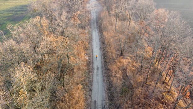 Woman in yellow jacket standing with a bicycle on black dirt road between trees and agricultural fields on sunny autumn day. Woman on bicycle on dirt road on dirt road. Top view. Travel tourism