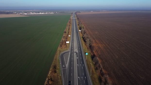 Beautiful panoramic landscape. Black paved highway with white markings,many driving passing cars, large agricultural fields with green agricultural crops and plowed black soil on a sunny autumn day.