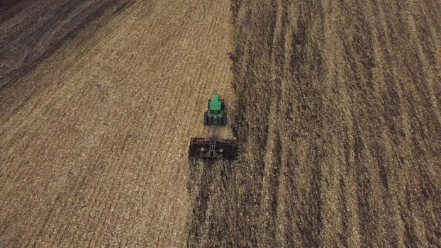Tractor plowing the ground. Flying over green tractor that plows up ground in yellow field after harvesting wheat on autumn day. Tractor digging land. Agricultural work on field. Aerial drone view