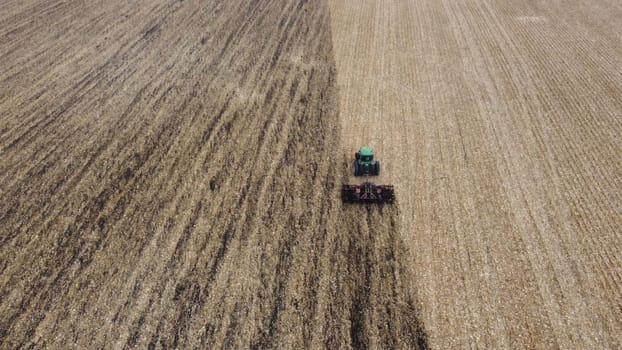 Tractor plowing the ground. Flying over green tractor that plows up ground in yellow field after harvesting wheat on autumn day. Tractor digging land. Agricultural work on field. Aerial drone view