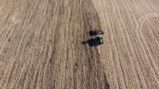 Tractor plowing the ground. Flying over green tractor that plows up ground in yellow field after harvesting wheat on autumn sunny day. Tractor digging land. Agricultural field work. Aerial drone view