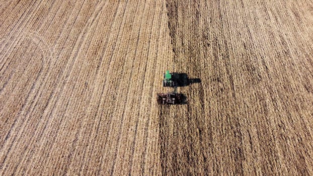 Tractor plowing the ground. Flying over green tractor that plows up ground in yellow field after harvesting wheat on autumn sunny day. Tractor digging land. Agricultural field work. Aerial drone view