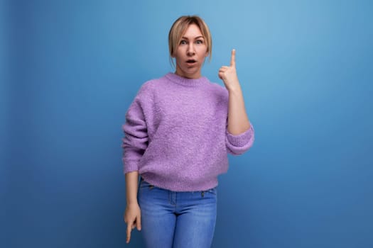 surprised pleasant blond young woman in purple sweater shows hand up on blue background with copy space.