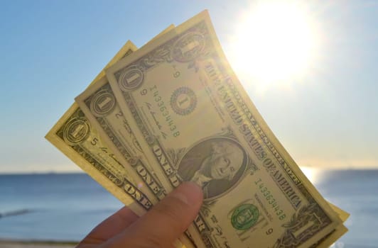 Person holding in hand paper bills of dollars with face value of one dollar on background of brightly shining sun, blue sky and calm blue sea. One-dollar paper banknotes on backdrop of blue sea sky