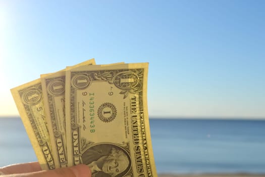 Man holding three one-dollar banknotes in his hand against backdrop of the clean blue sea and blue sky on sunny summer day. The concept of money, finance, monetary, travel, tourism, vacation, rest